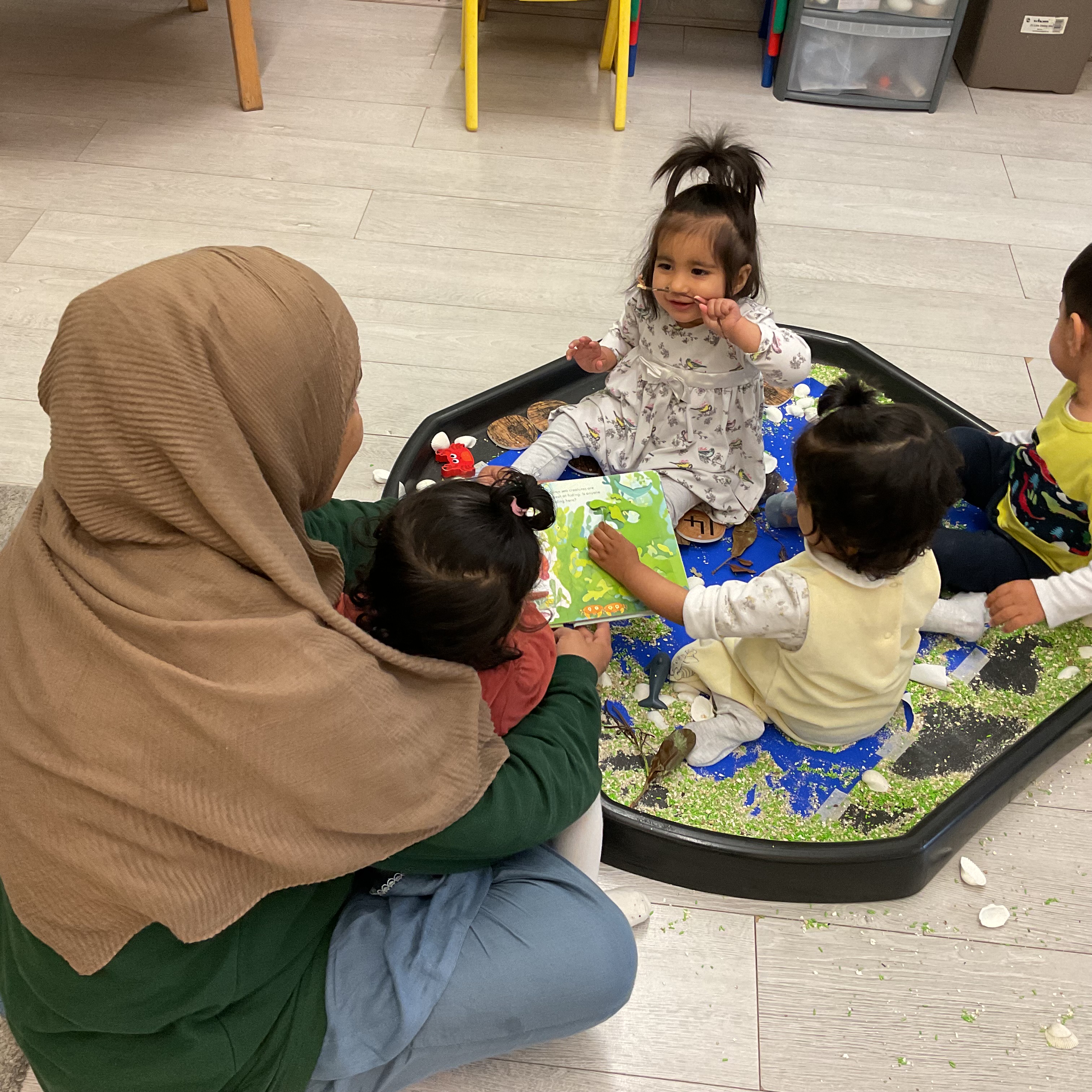 A teacher reading a picture book with children around a sensory tray at Apple Tree Montessori Nursery